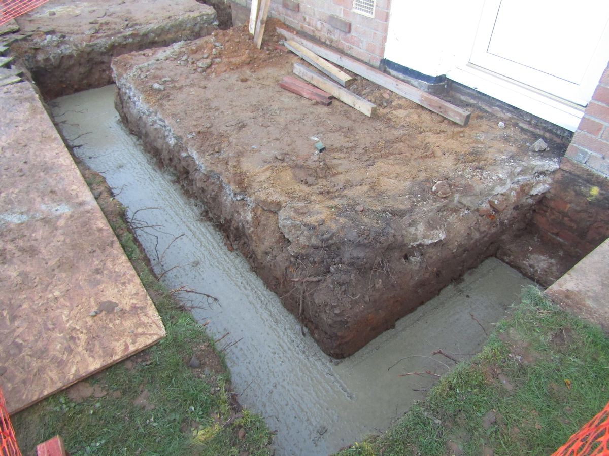 Freshly poured concrete filling a strip footing trench for a house extension, with the excavated soil banked alongside