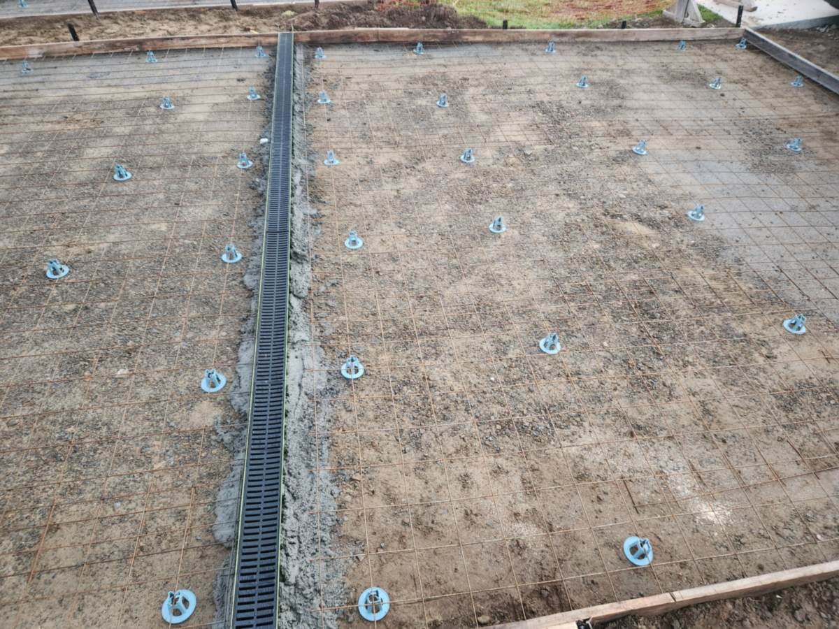Steel reinforcing mesh sitting on spacers inside timber formwork, with a channel drain along one edge, ready for a concrete garage base to be poured